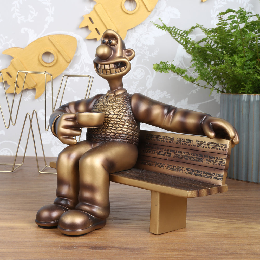 Bronze figurine of Wallace, sat on a bench grinning with a cup of tea in his hand. A tribute to the voice actor, the bench is engraved with facts about Peter Sallis. The background features wooden rockets and fern plant. 