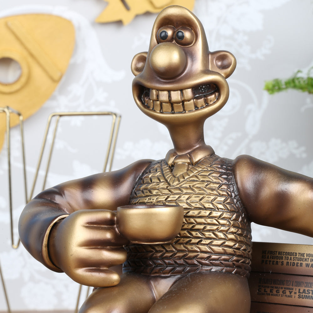 Bronze figurine of Wallace, sat on a bench grinning with a cup of tea in his hand. A tribute to the voice actor, the bench is engraved with facts about Peter Sallis. The background features wooden rockets and fern plant. 