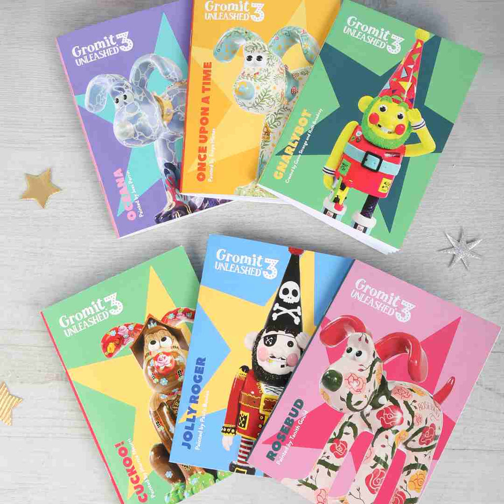 Collection of colourful books featuring Gromit, Wallace, Norbot and Feathers McGraw sculptures from the Gromit Unleashed 3 trail on a light grey wooden background