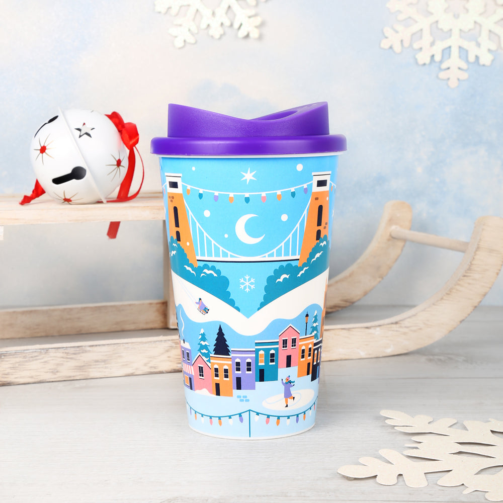 A travel coffee cup featuring a Bristol winter Christmas design with Bristol landmarks in a snowy scene
