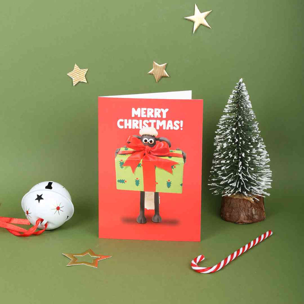Christmas card with Shaun the Sheep holding a gift, surrounded by festive decorations on a green background.