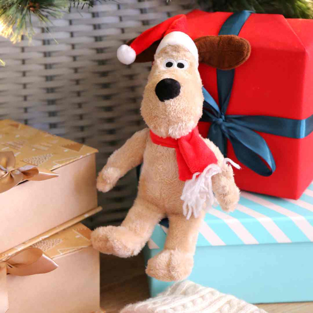 Small Gromit Soft toy with a santa hat and scarf sat on gifts under a Christmas tree.