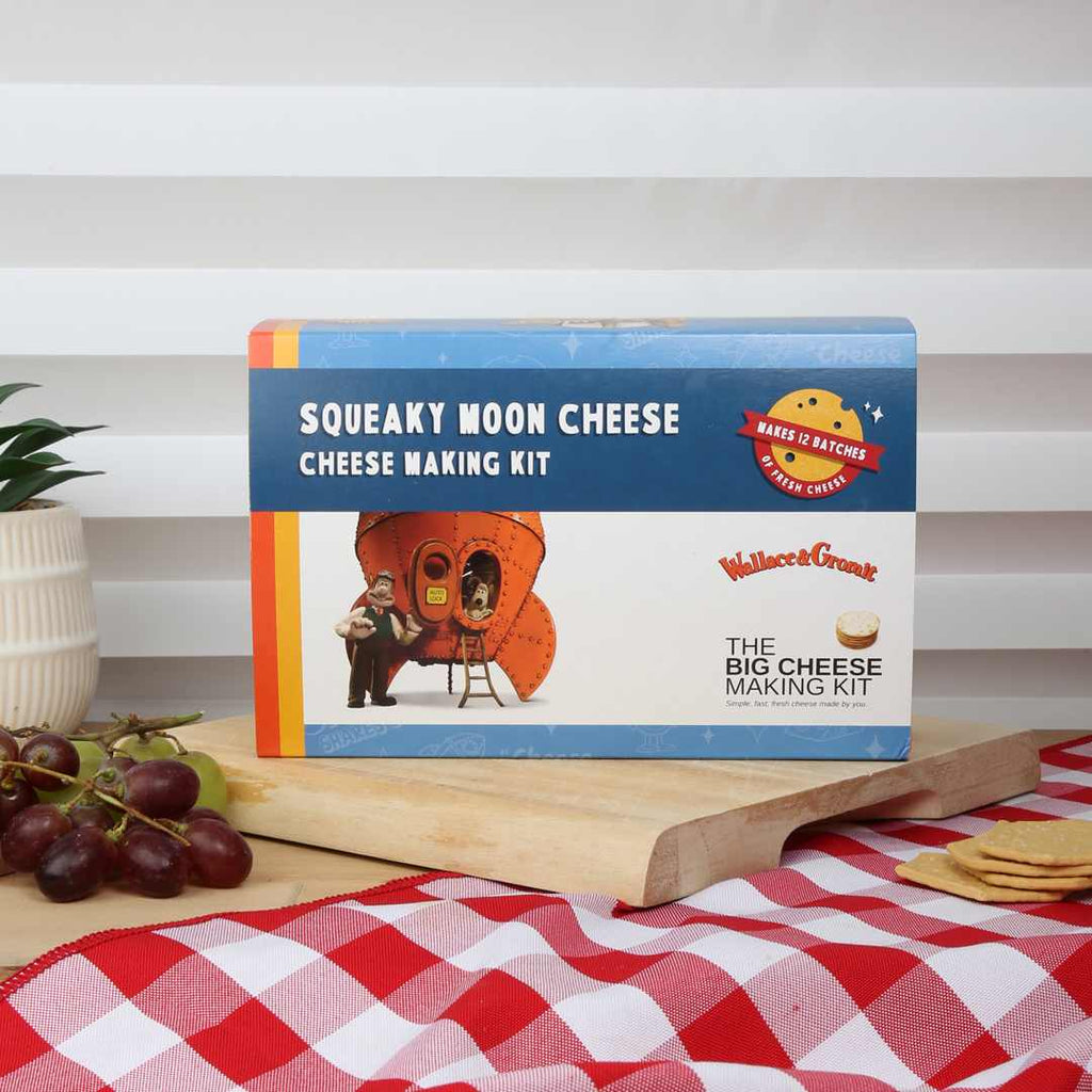 Wallace and Gromit themed squeaky Moon Cheese cheese making kit on a wooden board with grapes and cracket on a checkered tablecloth.rs