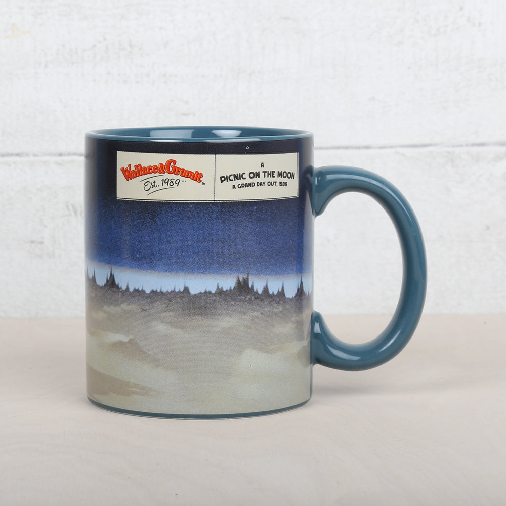 Mug with a blue handle, featuring a scene from Aardman's 'A Grand Day Out film'. Pictures Wallace holding a picnic basket and kicking a ball with Gromit by his side. The iconic orange rocket is in the background. 